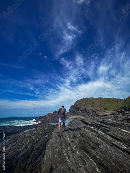 Obraz A young man walks on the rocks along the shore of a beach in the town of Búzios, the main tourist destination in the lakes region of Rio de Janeiro.