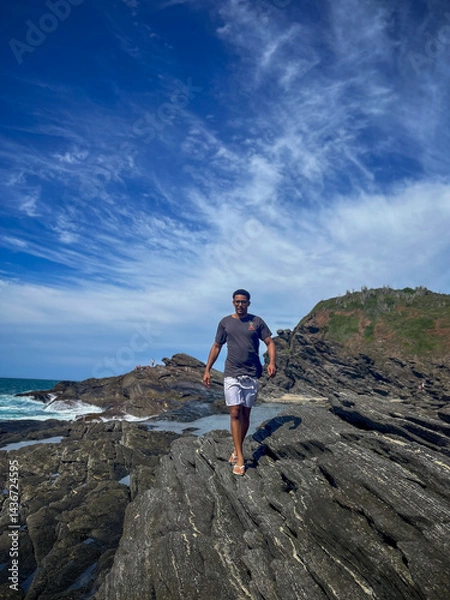 Obraz A young man walks on the rocks along the shore of a beach in the town of Búzios, the main tourist destination in the lakes region of Rio de Janeiro.