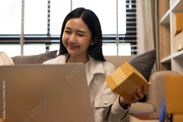 Fototapeta A woman is holding two boxes and smiling. She is sitting in front of a laptop. The boxes are brown and appear to be gifts. The woman seems happy and excited about the boxes
