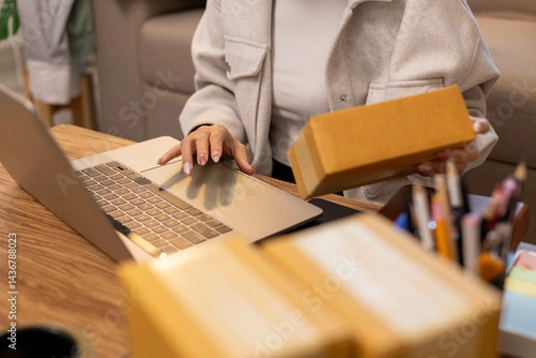 Fototapeta A woman is holding two boxes and smiling. She is sitting in front of a laptop. The boxes are brown and appear to be gifts. The woman seems happy and excited about the boxes
