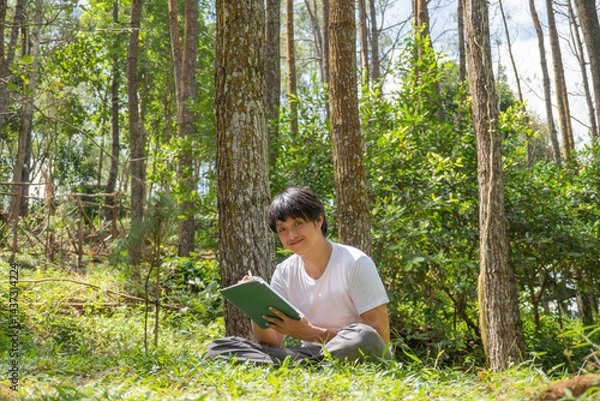 Obraz A young asian man wearing a white t-shirt and gray pants is drawing using a digital tablet while sitting on the grass in a lush pine forest in the morning. Remote work concept