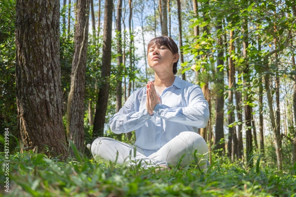 Obraz A young asian woman wearing a long-sleeve collared shirt and denim jeans pants is meditating and sitting on the grass in a lush pine forest in the morning.