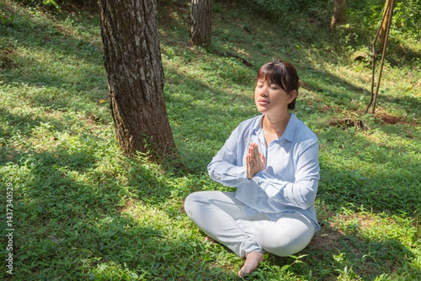 Obraz A young asian woman wearing a long-sleeve collared shirt and denim jeans pants is meditating and sitting on the grass in a lush pine forest in the morning.