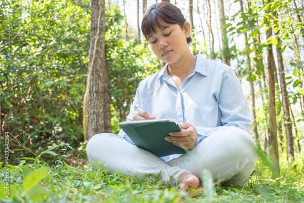 Obraz A young asian woman wearing a long-sleeve collared shirt and denim jeans pants is drawing using a digital tablet while sitting on the grass in a lush pine forest in the morning. Remote work concept