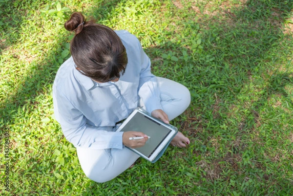 Obraz A young asian woman wearing a long-sleeve collared shirt and denim jeans pants is drawing using a digital tablet while sitting on the grass in a lush pine forest in the morning. Remote work concept