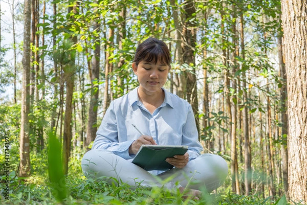 Obraz A young asian woman wearing a long-sleeve collared shirt and denim jeans pants is drawing using a digital tablet while sitting on the grass in a lush pine forest in the morning. Remote work concept
