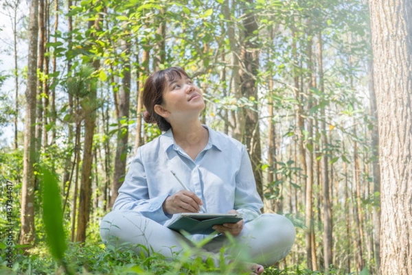 Obraz A young asian woman wearing a long-sleeve collared shirt and denim jeans pants is drawing using a digital tablet while sitting on the grass in a lush pine forest in the morning. Remote work concept