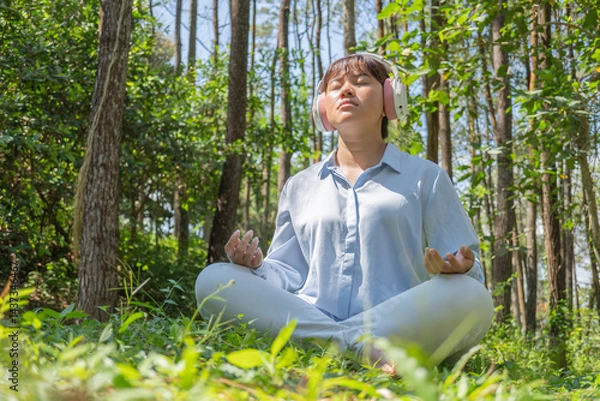 Obraz A young asian woman wearing a long-sleeve collared shirt and denim pants is meditating while listening to calming music through earphones, sitting on the grass in a lush pine forest in the morning.