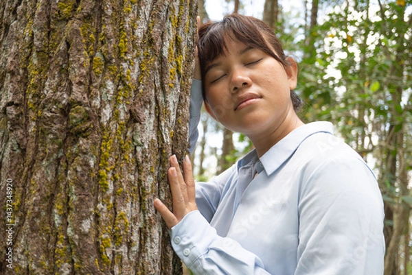 Obraz A Happy young asian woman wearing a long-sleeve collared shirt is hugging large pine tree trunk in a lush pine forest.