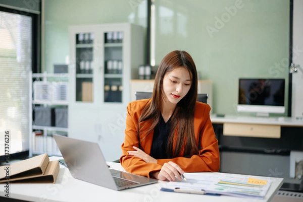 Fototapeta Asian woman sitting at a desk using a laptop computer Navigating Finance and Marketing