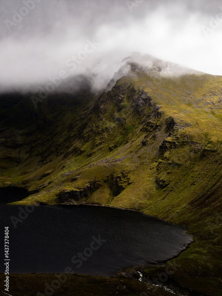 Obraz Misty mountain peak dramatically overlooks a dark, still lake, creating a serene yet powerful landscape. The contrast between the light and dark is breathtaking.