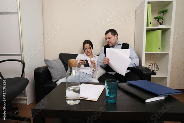 Fototapeta Man and woman seated on a black leather couch in a modern office, focused on reviewing documents together, representing team discussion, business strategy, or paperwork review scenarios.