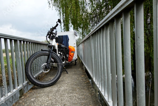 Fototapeta Electric bicycle towing small caravan crossing narrow bridge in countryside