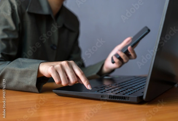 Fototapeta Woman in green blazer multitasking at office wooden desk. Female using laptop with one hand while holding smartphone in the other