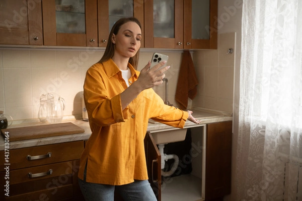 Fototapeta Homeowner woman is making a video call with her smartphone to show a broken sink drain to a plumber, explaining the problem and asking for assistance