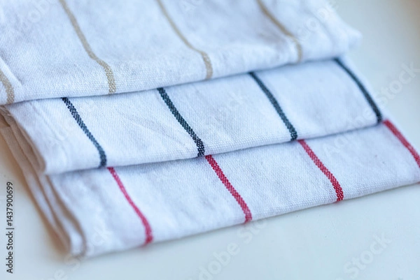 Obraz Neatly folded, three white tea towels with thin stripes in neutral tones of gold, dark blue, and red sit on a white surface.