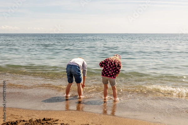 Obraz Two girls, fully dressed, playing with sand and waves and dipping their feet into the Mediterranean water, having a fun, joyful time. Children at the beach when it is too cold to swim