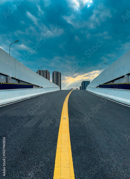 Fototapeta Wide highway stretches towards a cloudy sky in a modern urban environment at sunset