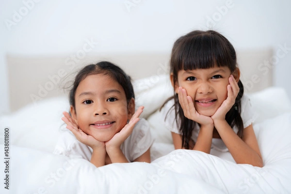 Fototapeta Two cute asian sisters lying on bed, resting heads on hands and smiling