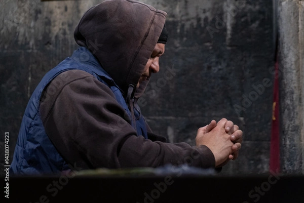 Obraz A man in a hoodie prays with folded hands inside a dark stone church. Deep moment of faith, meditation, and reflection in a historical religious place.