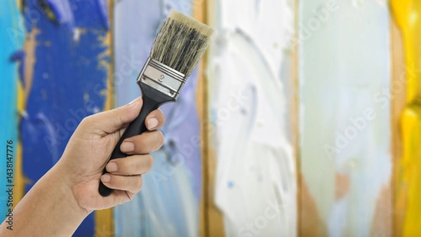Obraz Hand holding a paint brush against a colorful wooden background for creative projects