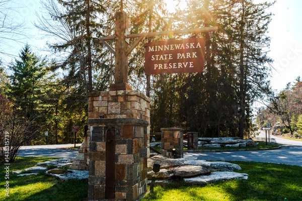 Obraz Kerhonkson, NY - US - Apr 23, 2025 Sunlight filters through tall pines at the entrance to Minnewaska State Park Preserve, where a rustic stone sign welcomes visitors to explore nature's beauty.