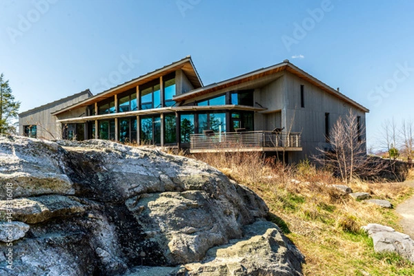 Fototapeta Kerhonkson, NY - US - Apr 23, 2025 Modern, wood-clad Minnewaska State Park welcome center with large glass windows sits beside stone outcrops, blending into the natural landscape under a clear sky.