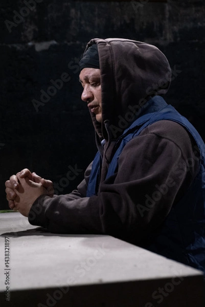 Obraz Vertical shot: An adult man wearing a hoodie prays in a church with clasped hands and closed eyes, asking God for blessings for his loved ones and peace for his country.