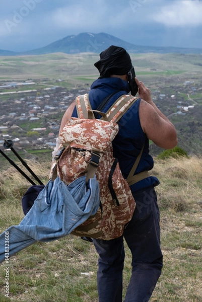 Fototapeta A rear view of an overweight male tourist wearing a headband and backpack, carefully descending a mountain trail while dark storm clouds gather above, fearing imminent rain.