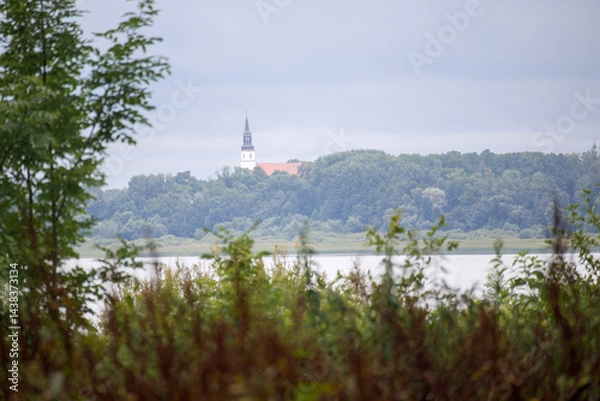 Fototapeta 
A landscape with a body of water in the foreground, behind which green trees spread out, and a building with a pointed tower, possibly a church, can be seen in the background.