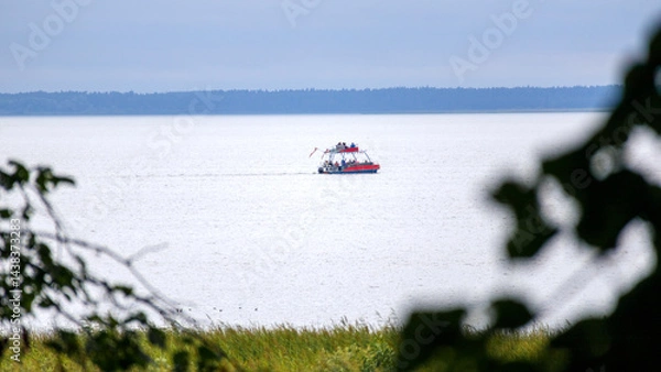Fototapeta 
A small, red boat with a white hull floats on a calm body of water in the distance, while blurred tree branches and grass are visible in the foreground.