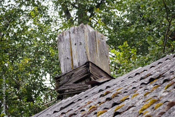 Fototapeta 
an old, damaged chimney with peeling plaster on a slate roof surrounded by green trees.
