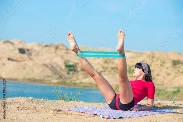 Fototapeta Slender athletic girl doing leg exercises with rubber expander on the sand of the beach near the water