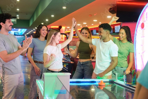 Obraz Group of friends giving high five while playing air hockey, celebrating victory in amusement park