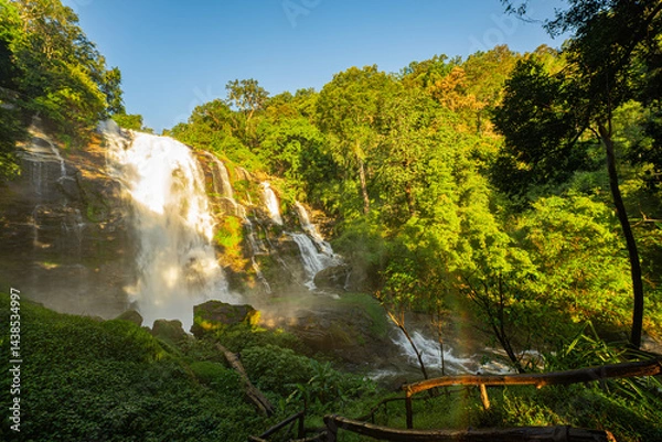 Fototapeta A scenic trail leads to a cascading waterfall surrounded by lush greenery, with a vibrant rainbow forming in the mist. This serene natural setting captures the harmony of water, light, 