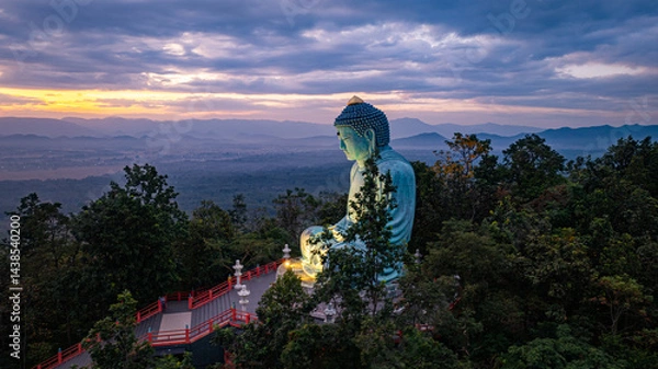 Fototapeta A serene aerial view of a large Buddha statue surrounded by lush forest, with a stunning sunrise casting vibrant hues over a misty mountainous landscape in the background. beautiful sky at sunrise.