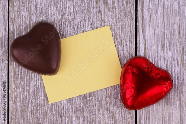 Obraz Two heart-shaped chocolates, one wrapped in red foil, placed on a rustic wooden background with a blank yellow card, perfect for Valentine's Day, love messages, or romantic celebrations.