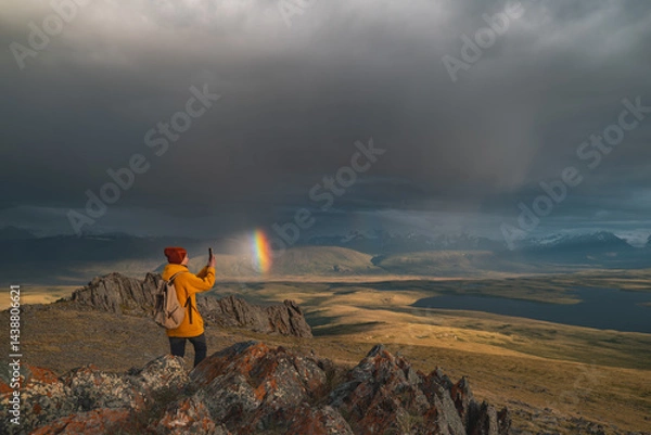 Fototapeta A traveler in bright clothes stands on the edge of a mountain as a rainbow appears in the sky. Golden hour in the mountains with a view of the rainbow and the valley illuminated by sunset.