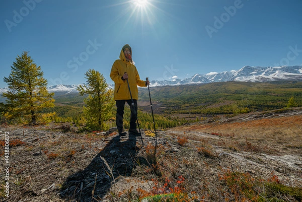 Fototapeta A man in bright clothes stands on the hill of a mountain, the sun is shining above his head, and behind him there are snowy mountains and a green forest. The action takes place in autumn in the mounta