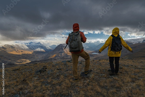 Fototapeta Urist travelers in bright clothes stand on a hill during the golden hour, in front of them there is a view of snowy mountains and valleys with lakes.