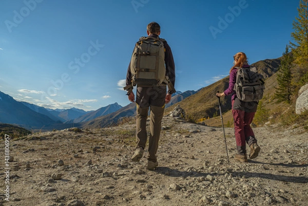 Fototapeta Two travelers in bright clothes with backpacks on their backs are walking along a mountain road to a slope with a magnificent view of a valley with a river and a forest.