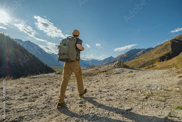 Fototapeta A human traveler with a backpack on his back walks along a mountain path, holding his hands by the straps of the backpack. In front of it there is a view of the mountains and the blue sky.