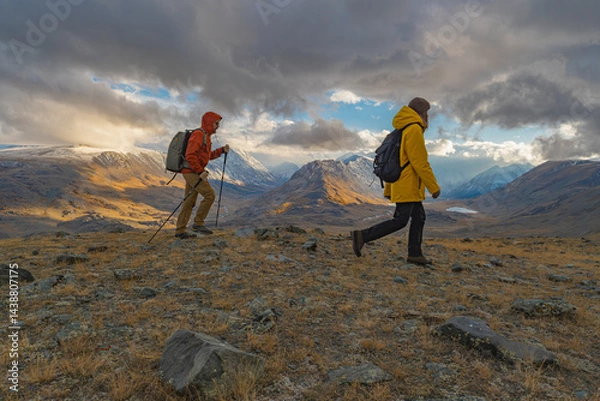 Fototapeta Travelers in bright clothes walk along the mountainside during the golden hour, overlooking the snowy mountains and the lake in the distance. Tourists with backpacks on their backs during sunset with 