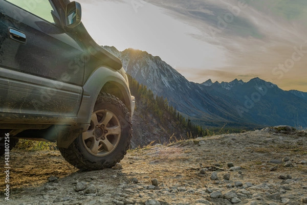 Fototapeta An off-road car stands on a mountainside during the golden hour, showing off off-road tires against the backdrop of a valley with mountains. Mud tires on the background of mountains.