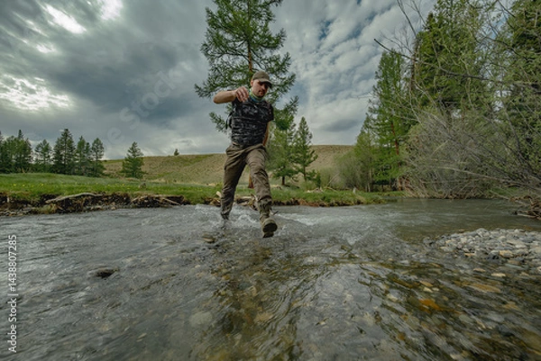 Fototapeta A traveler runs across a wide river against the background of a green forest. A man in boots runs through the water. Trekking clothes for travel.