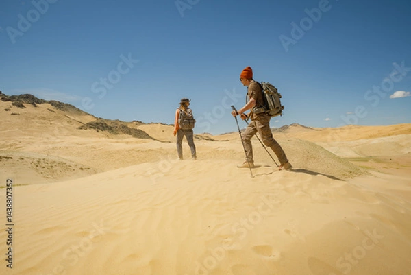 Fototapeta Two people with backpacks on their backs and in bright clothes are standing in the desert on a sand dune. Travelers stand on a dune with a view of the sky and small hills of plants.