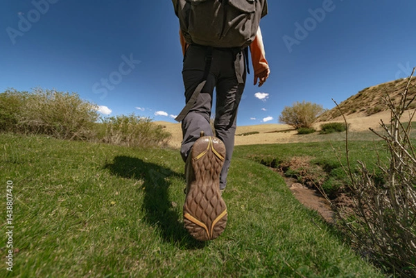 Fototapeta A traveler with a backpack on his back, walking through the green grass, defiantly showing the sole of his shoe. But the background is a sandy dune, bushes and a blue sky.