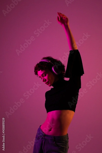 Fototapeta Cheerful young woman in wireless headphones feeling the rhythm, dancing with closed eyes in neon lights, against a red studio background.
