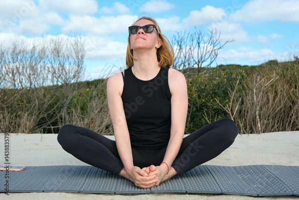 Obraz Woman practicing yoga outdoors, sitting in butterfly pose on a mat, enjoying fresh air, natural surroundings, and blue sky, embracing mindfulness and relaxation.