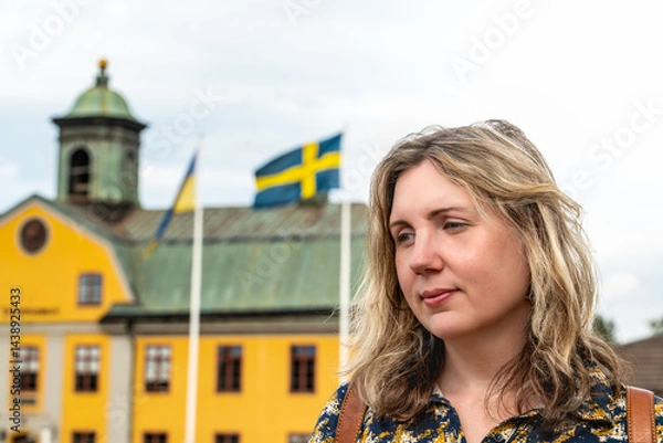 Fototapeta A woman poses thoughtfully with the Swedish and Ukrainian flags in view, set against a colorful building in Sweden during a sunny summer day